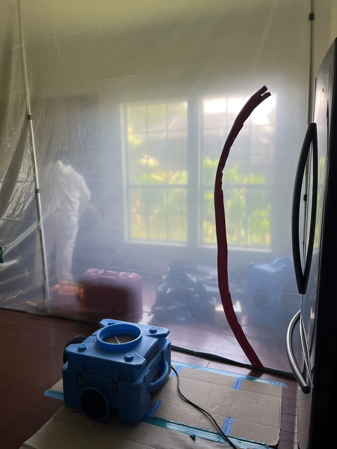 Technician in a protective suit working inside a sealed plastic containment room with a blue air scrubber in front