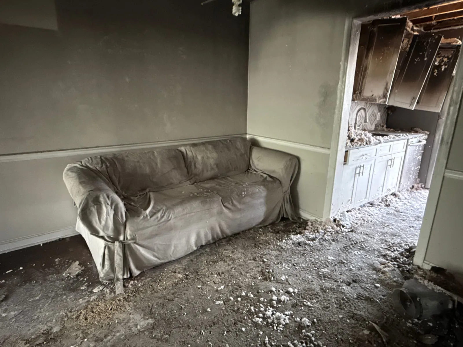 Living room with a soot-covered sofa and walls, burned kitchen cabinets through the doorway