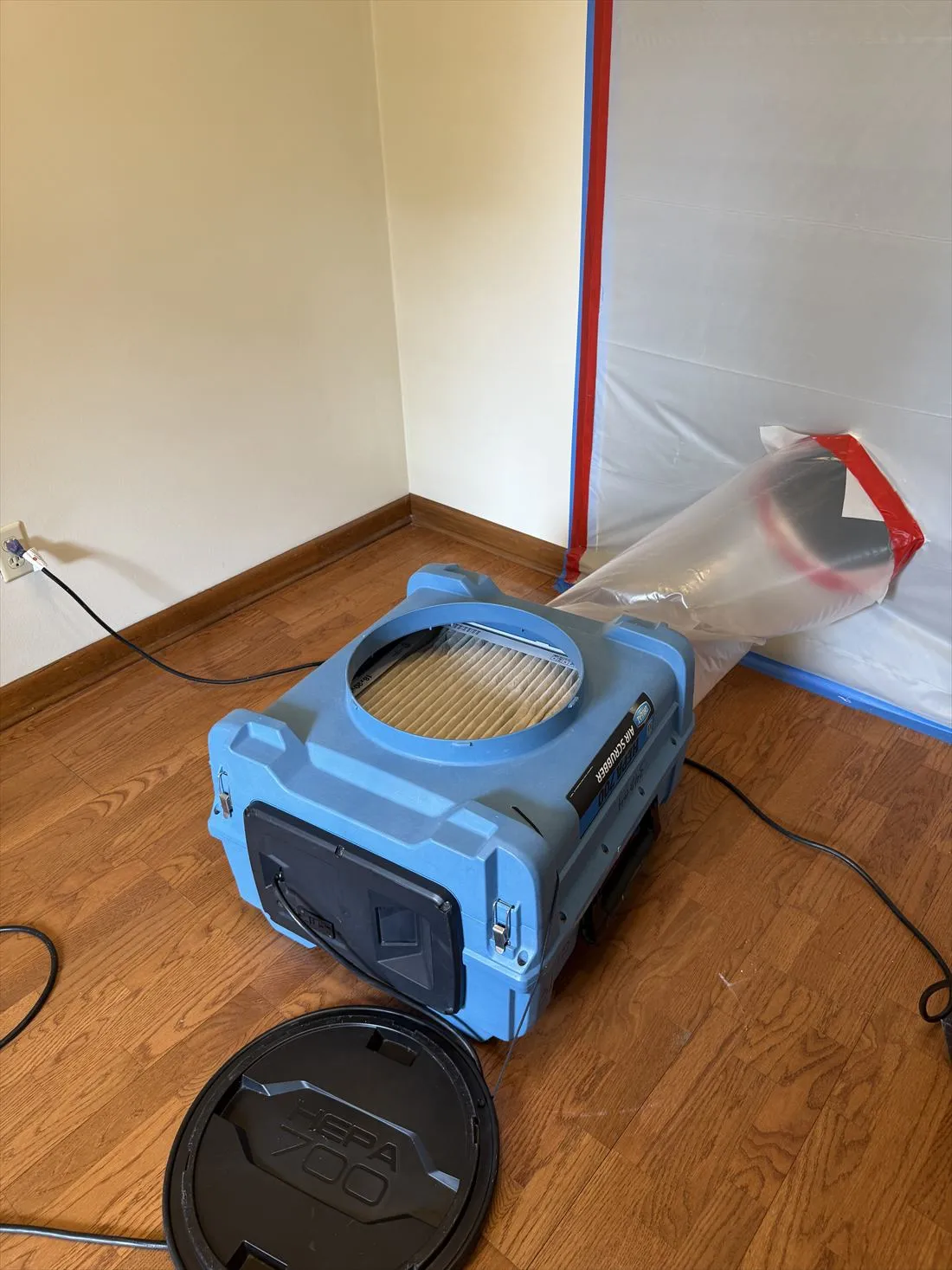 Blue HEPA air scrubber beside a sealed plastic containment wall in a room